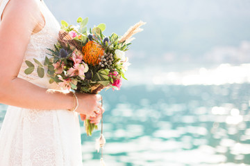 Bride with wedding bouquet is on beach near blue sea after ceremony. Bridal dress is white, lace,...