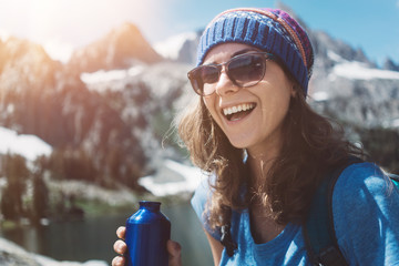 Portrait of handsome smiling hiking woman standing in stunning mountain wilderness with bottle near snowy lake - Powered by Adobe