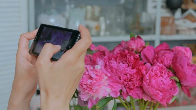 Professional floral artist, florist taking photo of her beautiful bouquet of pink peonies and carnations with smartphone. Woman's hands with mobile. Photography, technology and social media concept