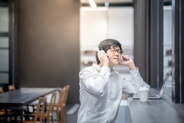 Young Asian man dressed in casual style using laptop computer while listening to music. Digital nomad working in co working space, modern IT lifestyle of freelance jobs