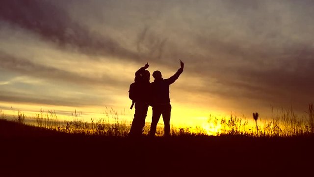 Silhouette of a couple of tourists with hiking backpacks ascending to the top of the mountain at sunset. Doing a selfie photo using a phone on top of a mountain. Slow motion. Travel concept.