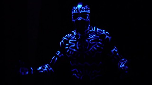 A crazy black man in neon patterns on the body dances and laughs in the dark, slow motion