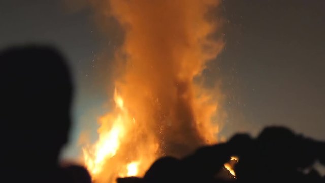 Close up of silhouettes of people watching large bonfire at night, some taking pictures. Focus on burning wood and flames