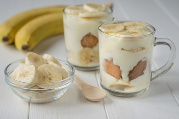 Two cups of banana pudding and chunks of banana in a glass bowl on a white table.