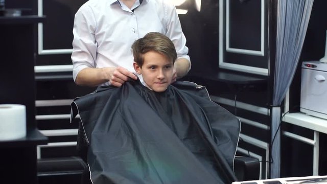 A male hairdresser puts on a young boy a protective cape before a haircut in the hairdresser.