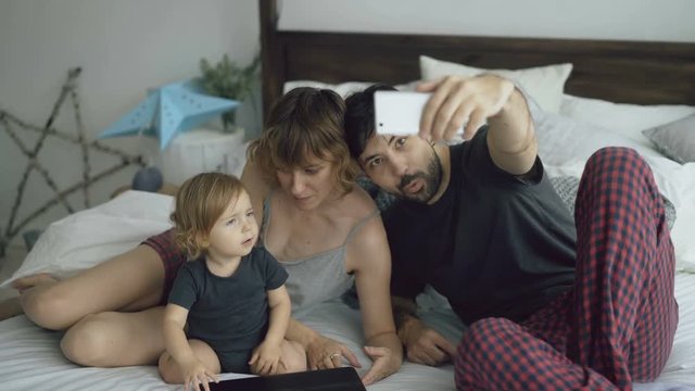 Young family with cute little girl taking selfie portrait on smartphone camera sitting in bed at home