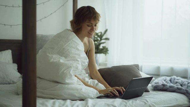 Cheerful young woman using her laptop computer sitting in bed at home