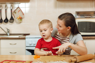 Fototapeta premium Little kid boy helps mother to cook Christmas ginger biscuit in light kitchen. Happy family mom 30-35 years and child 2-3 roll out dough and cut out cookies at home. Relationship and love concept