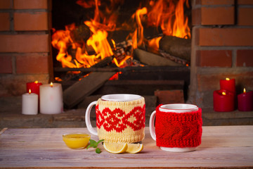 Hot drinks in mugs on wooden table beside cosy open fire place. Autumn or winter holidays concept, horizontal
