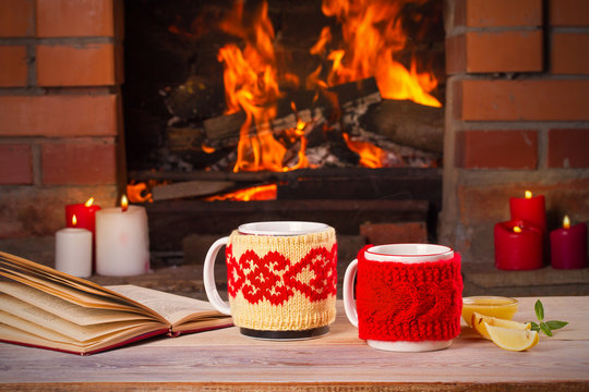 Hot drinks in mugs and book on wooden table beside cosy open fire place. Autumn or winter holidays concept, horizontal
