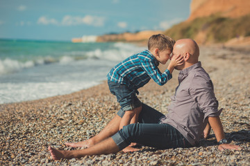 Touching appealing scene of father and son enjoy summer vacation together playing on stone beach wearing stylish shirt and fashion blue jeans both barefoot with adore landscape on background