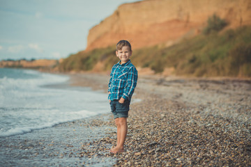 Fototapeta na wymiar Cute boy kid child wearing stylish shirt and blue jeans barefoot posing running on stone beach with gorgeous ocean sea landscape sand cliff cape