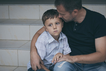Portrait of young sad little boy and father sitting outdoors at the day time.