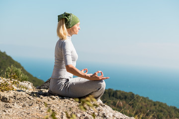 Fototapeta na wymiar Beautiful blond yoga woman sitting on the top of the mountain in lotus pose. Meditation on the edge with a scenic view of the landscape and the sea.