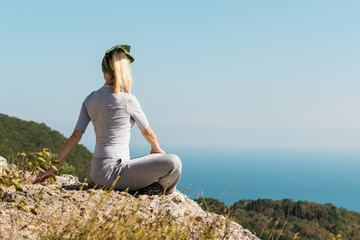 Naklejka na ściany i meble Beautiful blond yoga woman sitting on the top of the mountain in lotus pose. Meditation on the edge with a scenic view of the landscape and the sea.