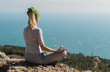 Beautiful blond yoga woman sitting on the top of the mountain in lotus pose. Meditation on the edge with a scenic view of the landscape and the sea.