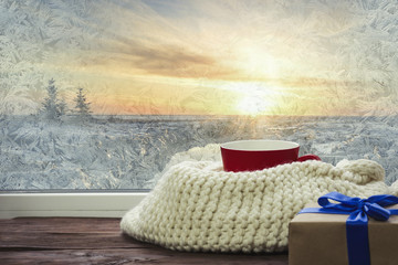 Fototapeta na wymiar On the background of a frozen window red cup, white knitted scarf and gift. Home cozy holidays background
