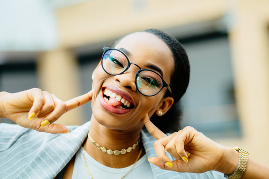 Goofy young teen african american woman student in eyeglasses making face with fingers pushing on cheecks and sticking out her tongue in a close up cropped head shot.