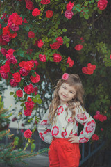 Cute baby girl with blond curly hairs and happy bright child eyes posing in central park close to huge bush of flowers red white pink roses wearing summer stylish clothes