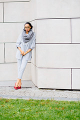 Verical full height of african business woman in glasses, gray scarf and suit, red shoes leaning on the beige wall of shopping center. Lifestyle, leisure and people concept.