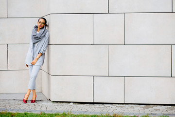 Horizontal full height of african business woman in glasses, gray scarf and suit, red shoes leaning on the beige wall of shopping center. Lifestyle, leisure and people concept.