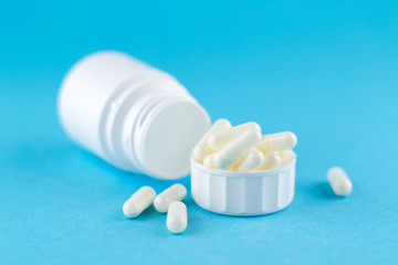 Close up white pill bottle with spilled out pills and capsules in cap on blue background with copy space. Focus on foreground, soft bokeh. Pharmacy drugstore concept