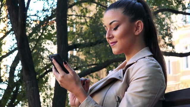 Young beautiful woman sits on the bench in park and works on the smartphone
