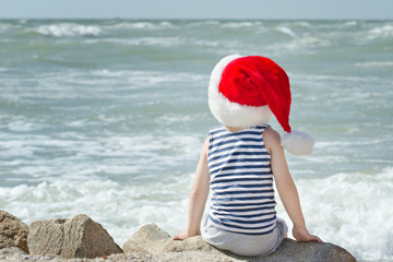 Boy in Santa hat sitting on the beach. Back view