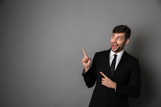 Overjoyed brunette man in classic suit pointing with two fingers, looking aside