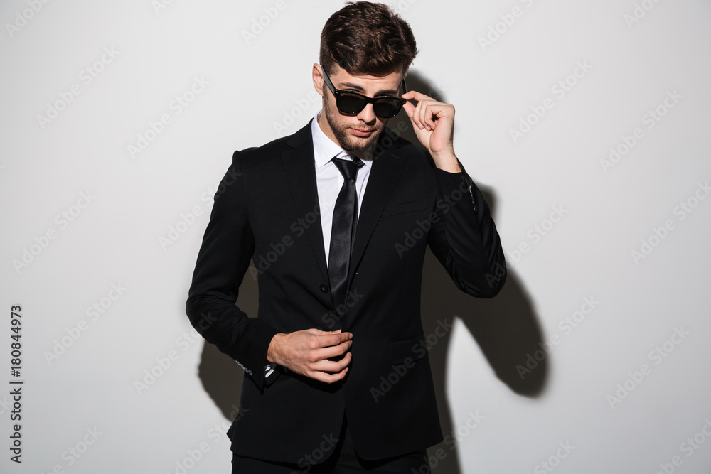 Poster close-up portrait of young bearded man in black suit touching his sunglasses, looking at camera