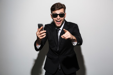 Close-up photo of young happy exited attractive man in sunglasses holding smartphone and pointing...