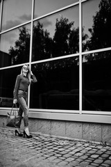 Portrait of a fabulous young woman in red blouse and jeans posing with her handbag and sunglasses outside the shopping mall on glass background. Black and white photo.