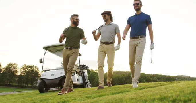 Three handsome caucasian men strolling with their clubs on the golf field near white golf car and talking. Outdoor