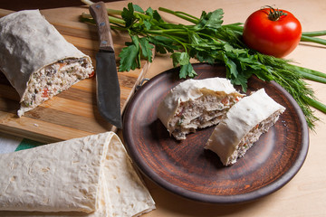 Two pieces of pita bread or lavash roll with cottage cheese or curd, chicken, tomatoes and herbs - dill, onion, parsley on brown clay plate..