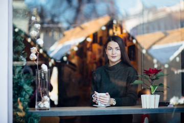 Beautiful brunette young woman in a cafe holding a cup of coffee or cocoa, seen through the window with buildings and lights reflections. She is looking away. Lifestyle concept.