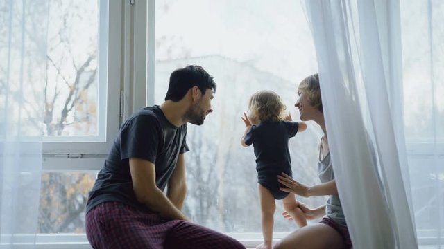 Happy family with young cute daughter sitting on windowsill playing and looking in window at home