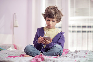 Little boy sitting on bed listening music on his smart phone.
