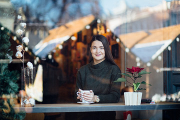 Beautiful brunette young woman in a cafe holding a cup of coffee or cocoa, seen through the window with buildings and lights reflections. She is looking away. Lifestyle concept.