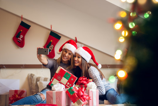 Two beautiful excited girlfriends with Santa hat sitting on the bed for Christmas holidays and taking a selfie with happiness.