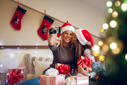Two adorable pretty girlfriends with Santa hat sitting on the bed for Christmas holidays one next to another and taking a selfie while having a friendly kiss on the cheek.