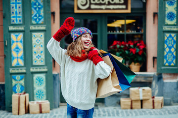 Young pretty woman near the shop with gifts smiling and saying hello in sunny winter day. Girl dressed in warm bright hat, red scarf and mittens. Happy holiday and freedom concept.
