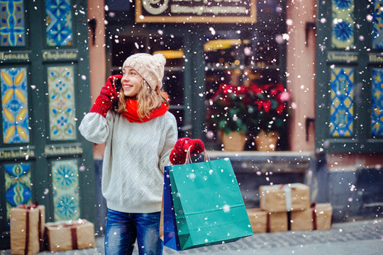 Beautiful Smiling Woman At The Street With Shopping Bags Talking On Mobile Phone. Attractive Girl Dressed In Warm White Hat, Red Scarf And Mittens. Magic Snowfall Effect.