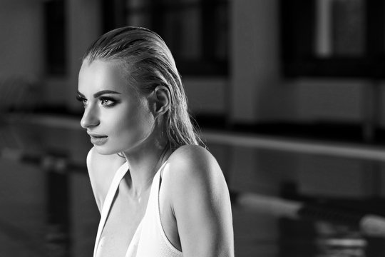 Seductive blonde woman with long wet hair dressed in white swimsuit with low-cut neckline standing in swimming pool and leaning on edge with her hands. Sexy female model emerging from blue water. - Powered by Adobe