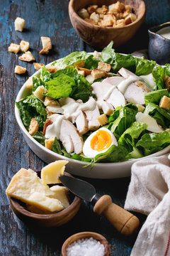 Classic Caesar salad with grilled chicken breast and half of egg in white ceramic plate. Served with ingredients above over old dark blue wooden background. Close up. Rustic style