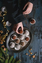 Child hands make homemade dark chocolate truffles with cocoa powder, coconut, walnuts as Christmas gift, put on vintage tray. Fir tree, Christmas decorations above on old wooden table. Top view.