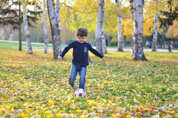 Boy kicks the ball in autumn park. Happy child play with ball