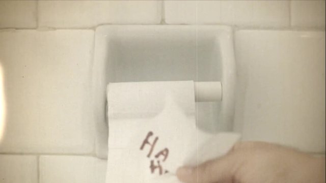 Fake 8mm amateur film: grabbing the last strip of paper from a toilet roll and discovering a mocking notice written in red ink.
