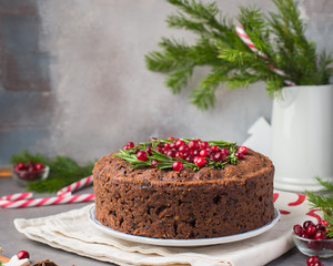 Traditional Christmas English pudding (fruit cake) with rosemary and cranberries on white plate on a concrete light background. Christmas decor of the table.  