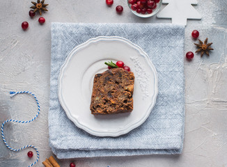 A piece of traditional Christmas English pudding (fruit cake) on white plate on a concrete light background. Christmas decor of the table. Funny napkins with reindeers Santa Claus.  Flat lay, top view