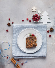 A piece of traditional Christmas English pudding (fruit cake) on white plate on a concrete light background. Christmas decor of the table. Funny napkins with reindeers Santa Claus.  Flat lay, top view